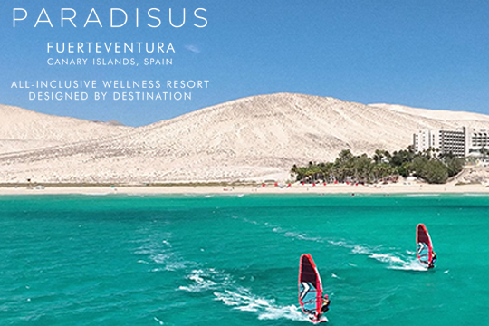 Zwei Surfer im Vordergrund, hinten Sandd&uuml;nnen und rechts ein Hotel